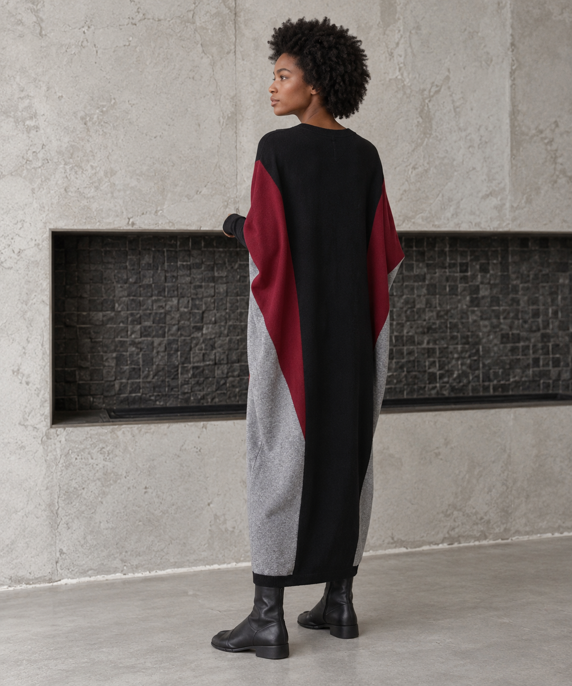 Rear three-quarter view of the oversized block knit midi dress, showing its black back, burgundy side panels and tapered grey hem.