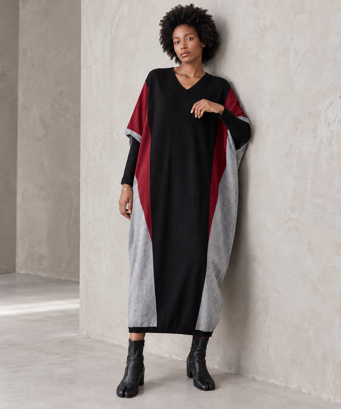 Model leaning against a concrete wall in the black oversized V-neck block knit midi dress, showing its loose drape and colour-block panels.