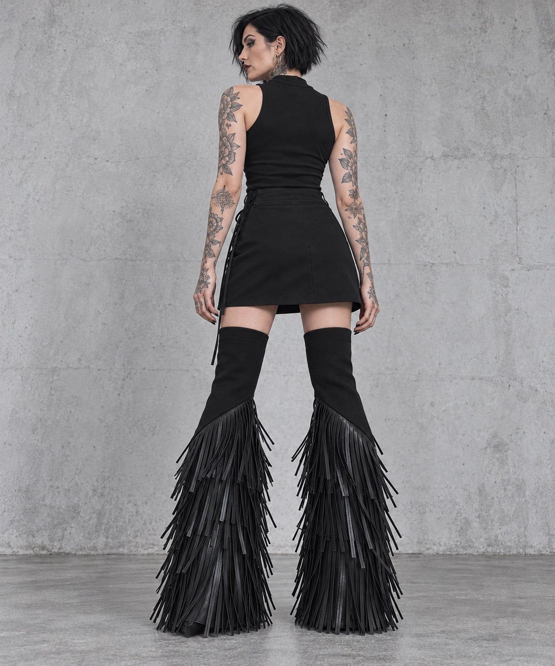 Back view of black vegan leather tassel flare leg warmers styled with a short black skirt.