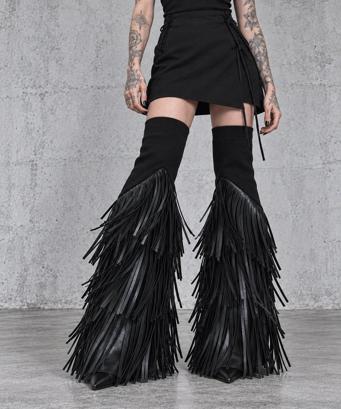 Close-up of black tassel flare leg warmers showing the separate upper cuffs and layered fringe over pointed black shoes.