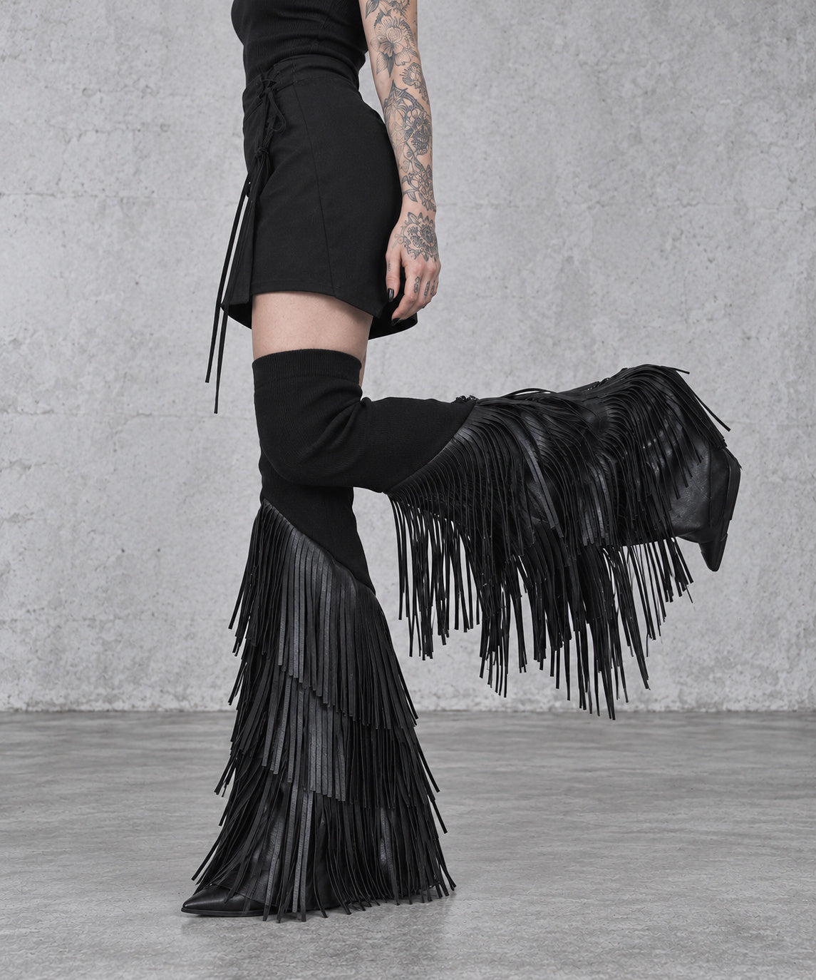 Side detail of black vegan leather tassel leg warmers with one knee bent, showing the hanging fringe and flared lower section.