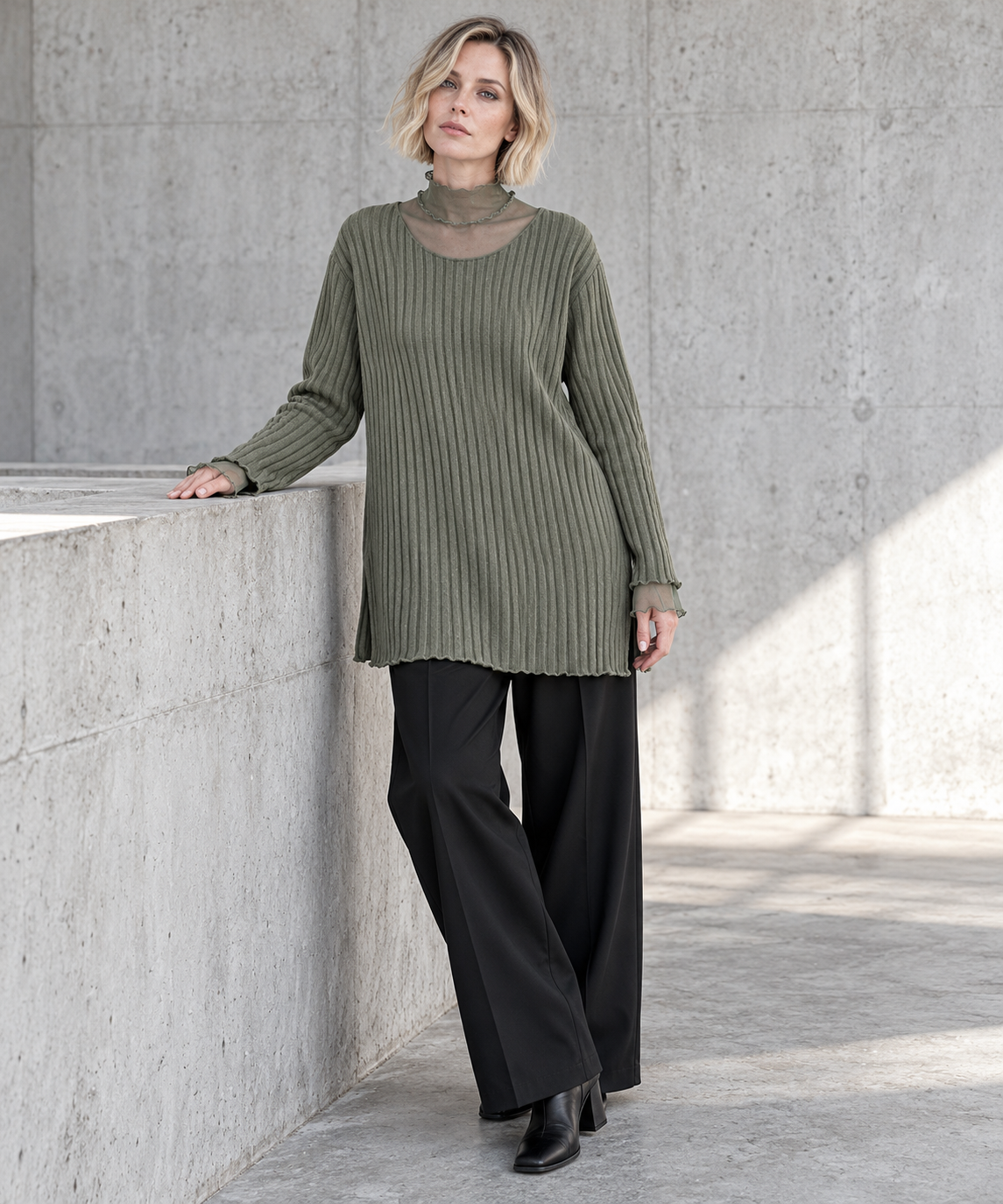 Olive Green Oversized Turtleneck Knit Sweater. Blonde model resting one hand on a concrete ledge, wearing the olive rib-knit sweater with black wide-leg trousers.